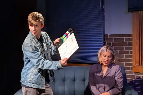 Asher holds up the clapboard while Erica waits on the news couch